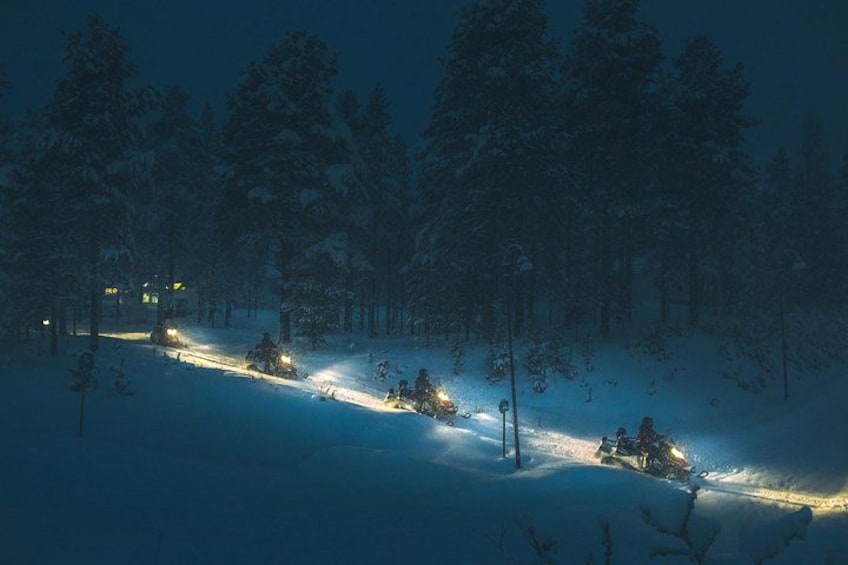 A snowy forest with a snowmobile trail and a lit building in the distance. 2