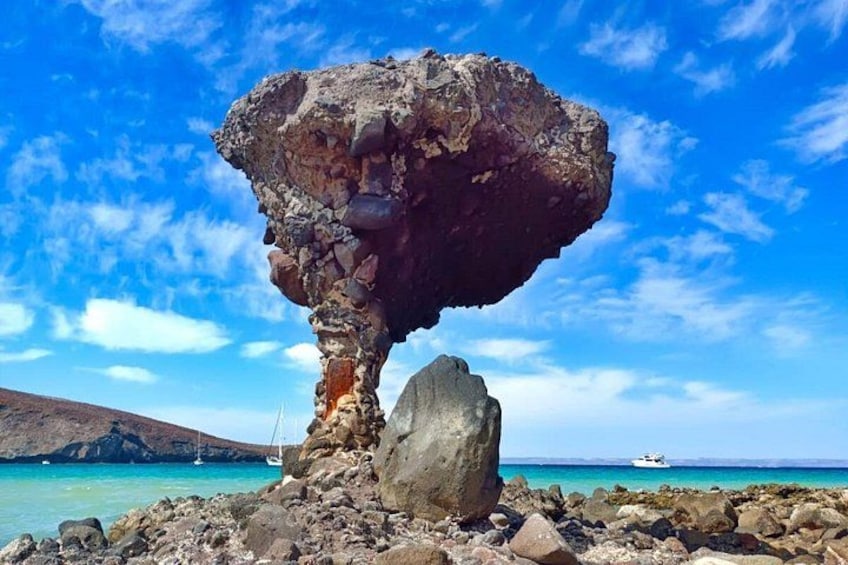 Formación rocosa singular con una roca equilibrada en la cima, cielo azul despejado y un barco a lo lejos. 1