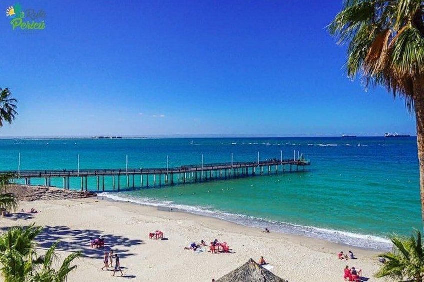 Una playa con un muelle que se adentra en el océano, palmeras y gente relajándose en la arena. 4