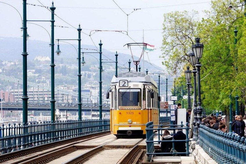A tram on tracks with overhead wires, a bridge, and a cityscape in the background. 3