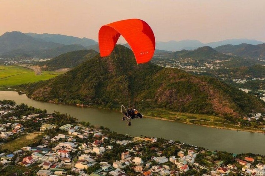 A paraglider flying over a river with a village below, surrounded by hills. 2