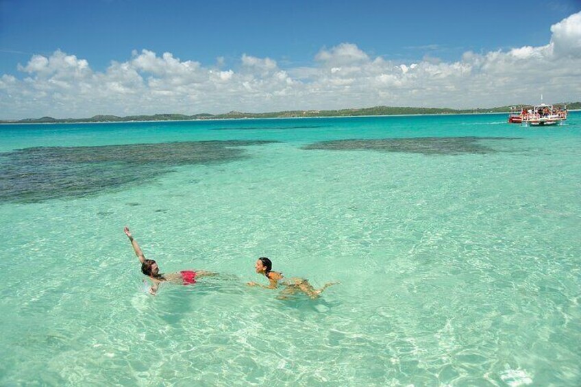 Two people swimming in clear turquoise water with a boat in the background. 1