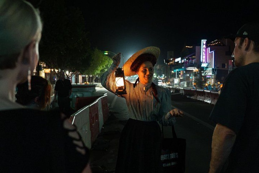 A person wearing a hat and holding a light, standing on a sidewalk at night with illuminated buildings in the background. 4
