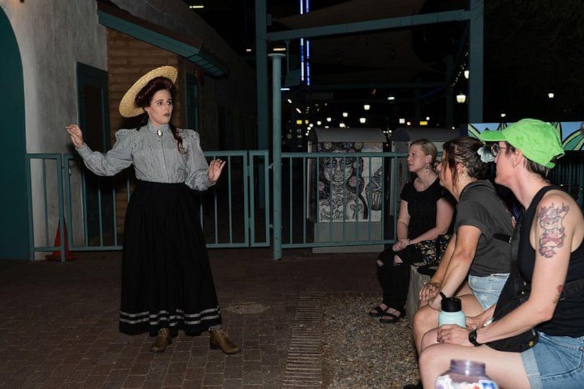 A person in a hat and dress standing with arms outstretched, and a group of people sitting on benches in an outdoor setting. 2