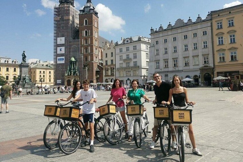 Een groep mensen op fietsen op een openbaar plein met historische gebouwen op de achtergrond. 1