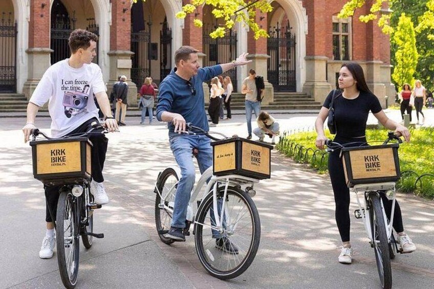 Three people riding bicycles with delivery boxes labeled 'KRB BIKE' in an outdoor setting with buildings in the background. 3