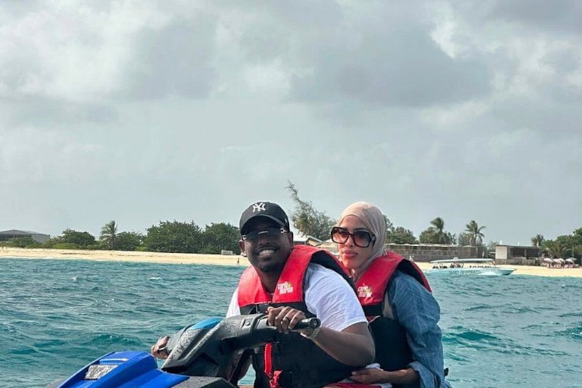 Two people on a jet ski, wearing life jackets, with a beach and palm trees in the background. 2
