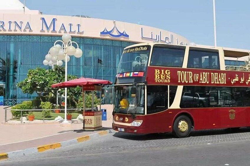 Ein roter Doppeldeckerbus mit der Aufschrift 'BIG BUS TOURS' und 'TOUR OF ABU DHABI', der vor einem Gebäude mit den Schildern 'MARINA MALL' und 'LIGAR' parkt. 5