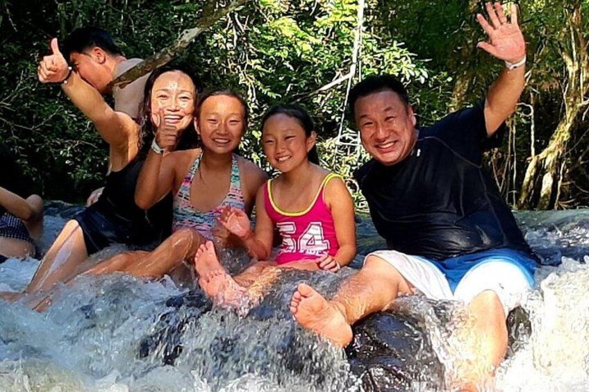 A group of people sitting on a rock in a flowing water stream surrounded by greenery. 1