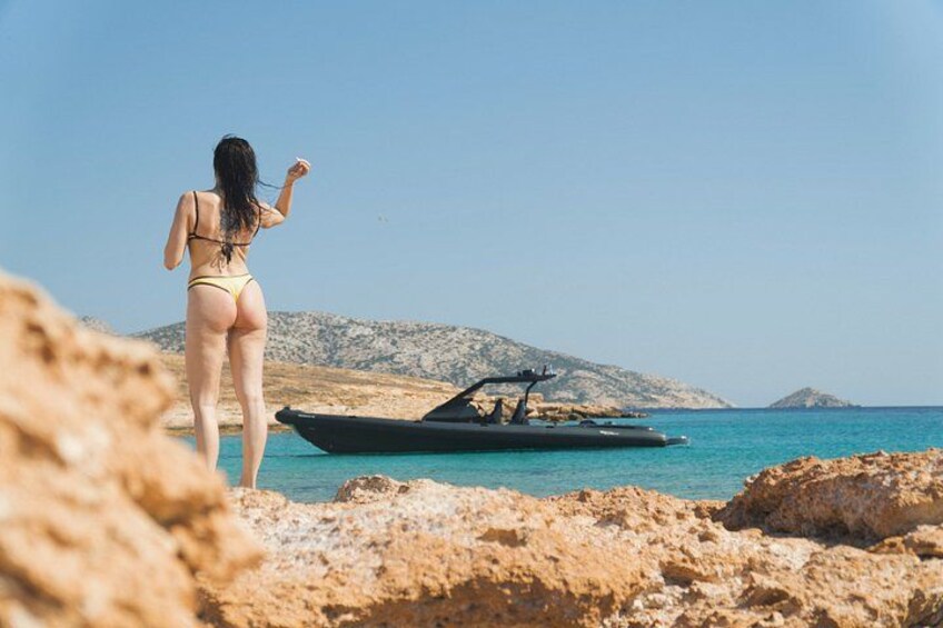 Eine Frau in einem Bikini geht über felsiges Gelände in Richtung eines Bootes auf ruhiger See, im Hintergrund eine hügelige Landschaft. 5