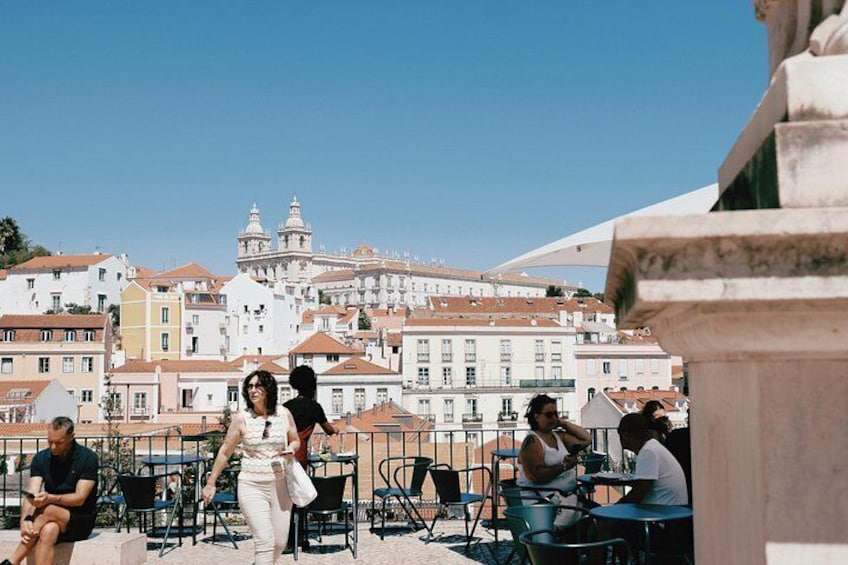 A group of people sitting at outdoor tables on a terrace with a cityscape in the background. 4