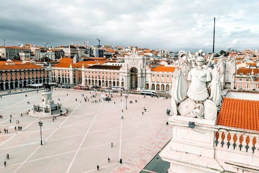 A plaza with a large statue, buildings with red roofs, and a fountain. 1