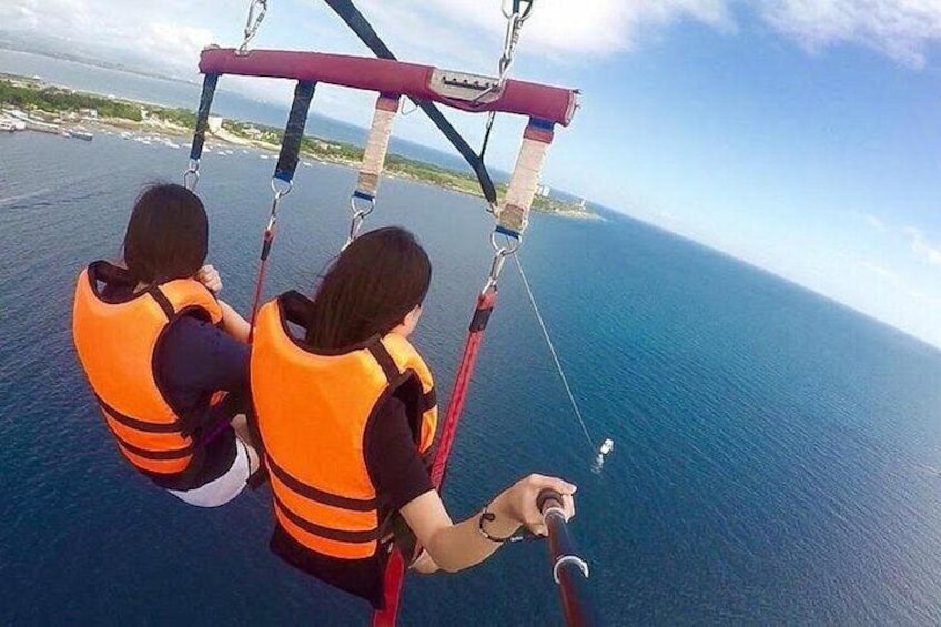 Two people in orange life jackets attached to a red harness system, suspended over a body of water. 2
