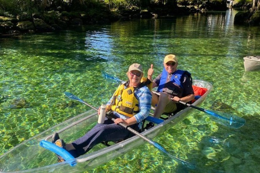 Two people in a transparent kayak on a clear river surrounded by greenery. 4