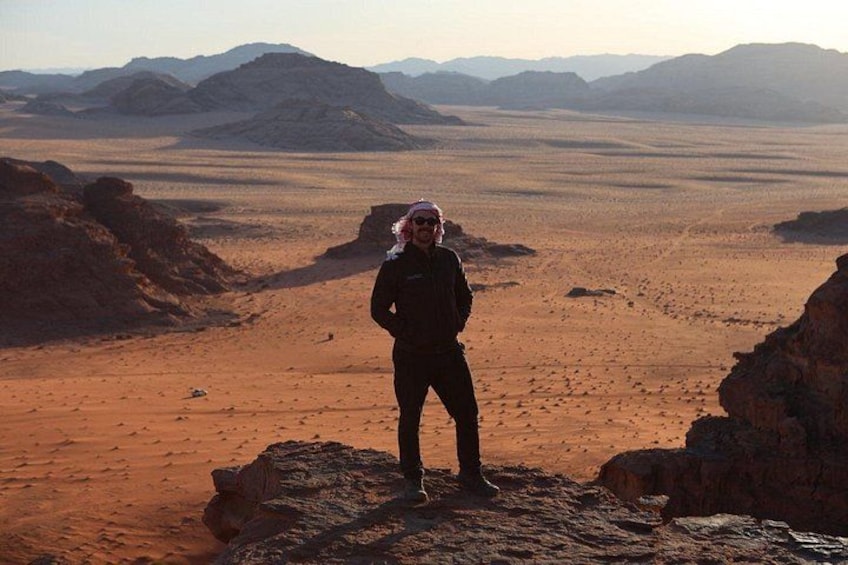A person standing on a rocky outcrop in a desert landscape with mountains in the background. 1
