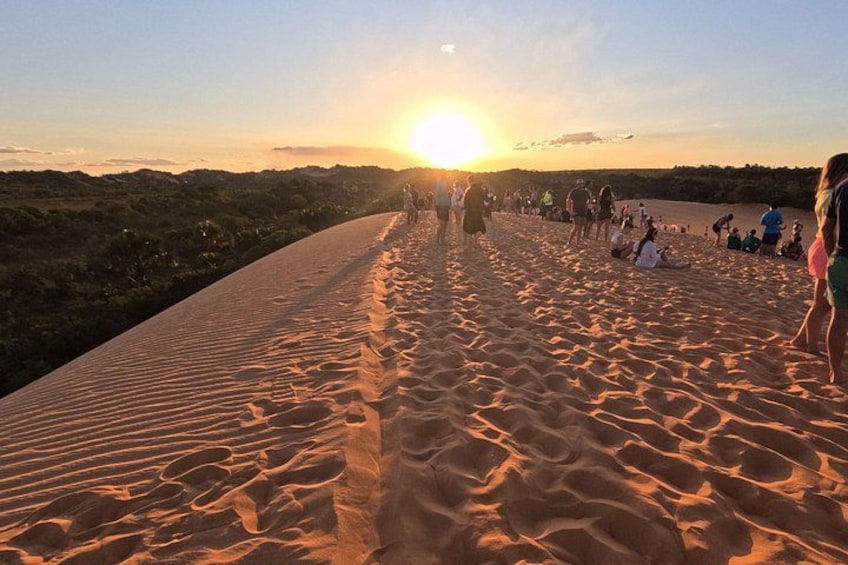 A sandy beach with people walking and sitting, and the sun setting in the background. 2