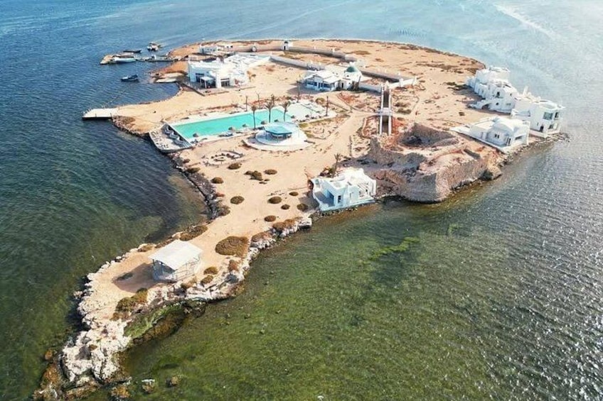 An aerial view of a coastal island with white buildings, a swimming pool, and clear blue waters. 5