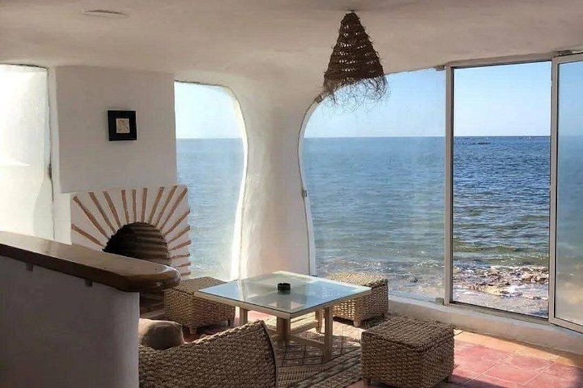 A room with a glass table and chairs, a woven chair, a hanging light fixture, and a view of the ocean. 2