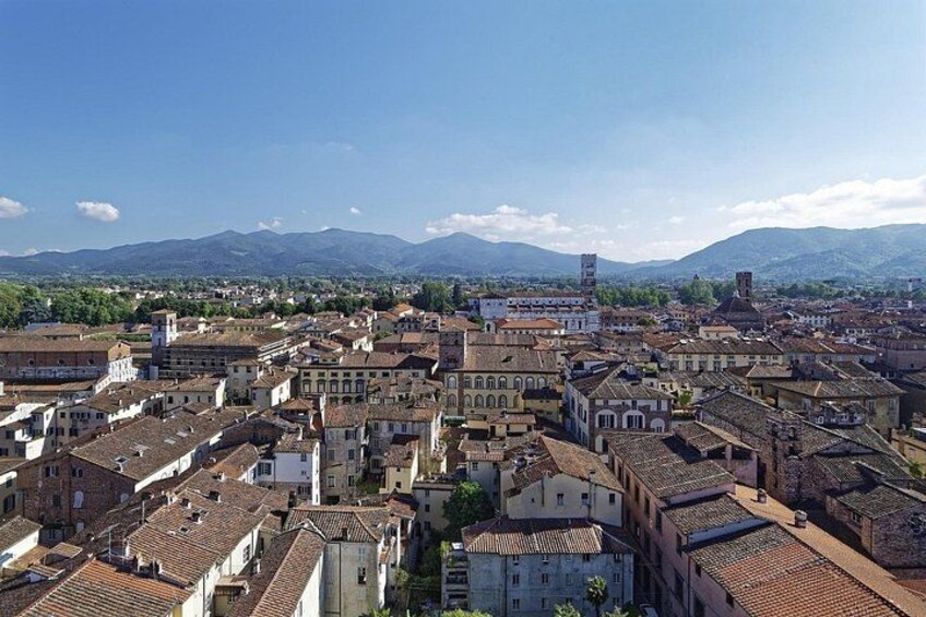 Une ville historique aux bâtiments aux toits de tuiles rouges, avec un clocher d’église imposant et des montagnes en arrière-plan. 1