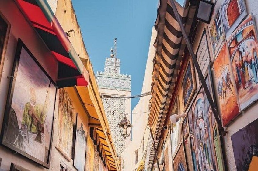 A narrow street with buildings on either side, a tower with a spire in the distance, and a clear blue sky. 4