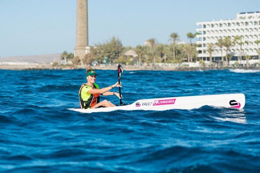 Una persona practicando windsurf en una tabla con un logotipo, en un cuerpo de agua con un edificio y palmeras al fondo. 1