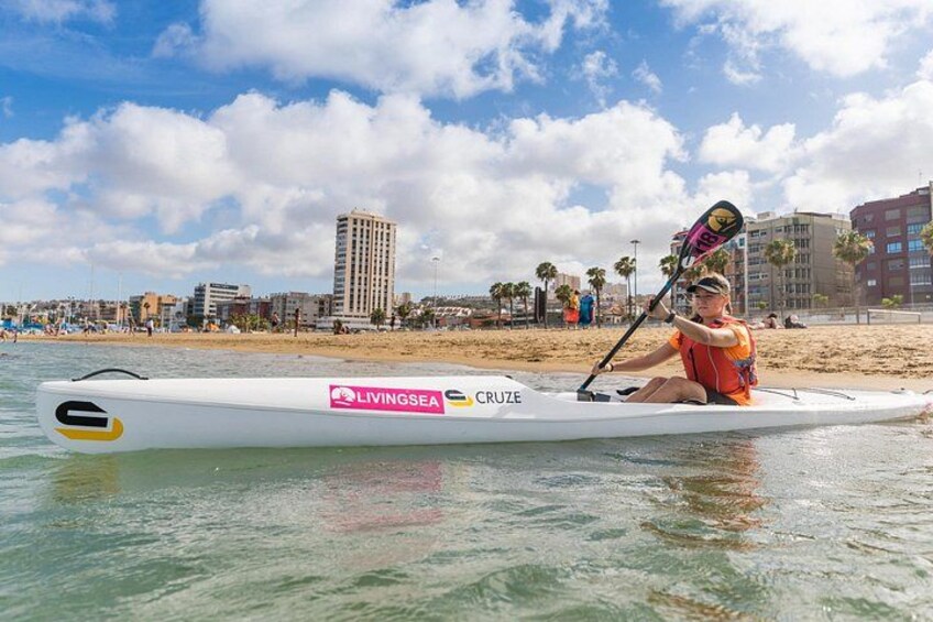 Una persona rema en una canoa blanca en la playa, con un paisaje urbano al fondo. 2