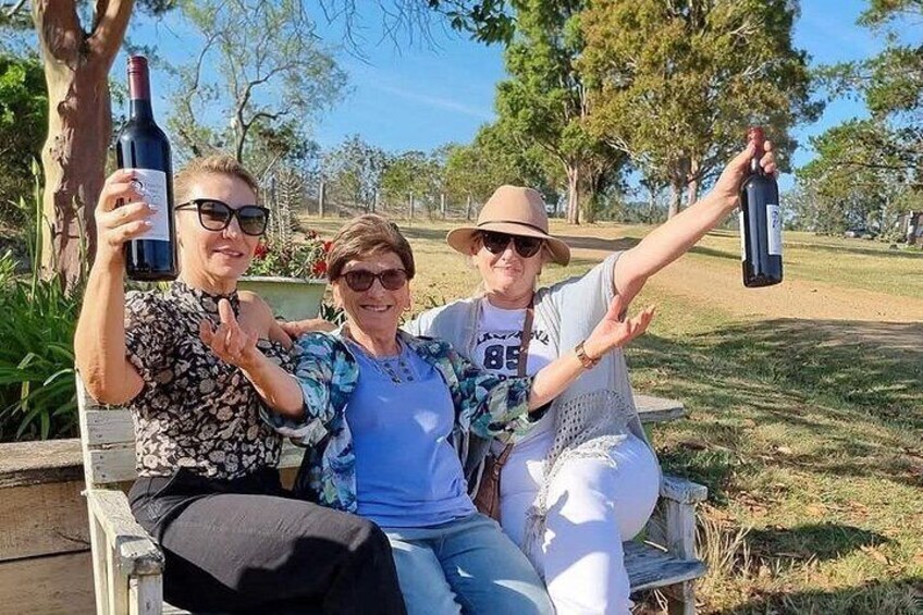 Three people sitting on a bench outdoors, holding wine bottles. 2