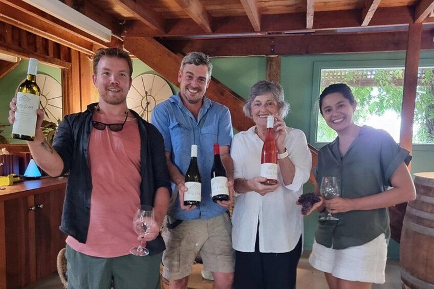 Four people holding wine bottles and glasses in a covered outdoor area with wooden beams and a green wall. 1