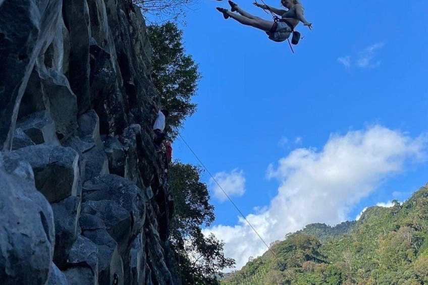 A person bungee jumping off a rocky cliff with a clear sky and forested mountains in the background. 2