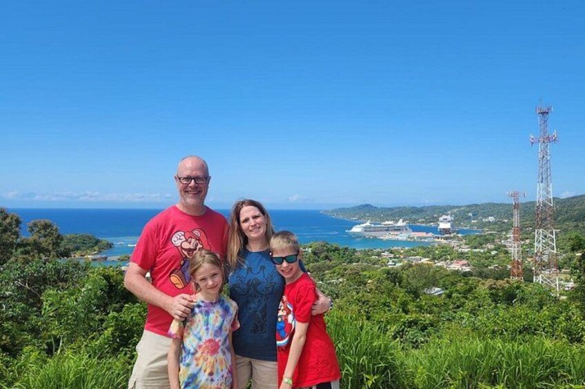 A family of four standing on a grassy hill with a view of the ocean and a tall communication tower in the background. 4