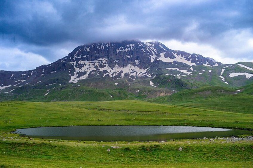 A mountain with snow patches, a green valley, and a small lake. 3
