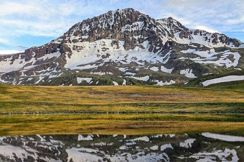 A mountain with snow patches, a grassy field, and a reflective water surface. 5