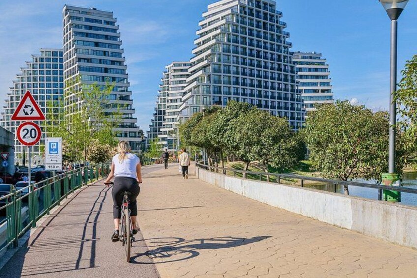 Een verhard pad met een fietser, een rivier en moderne gebouwen op de achtergrond. 1
