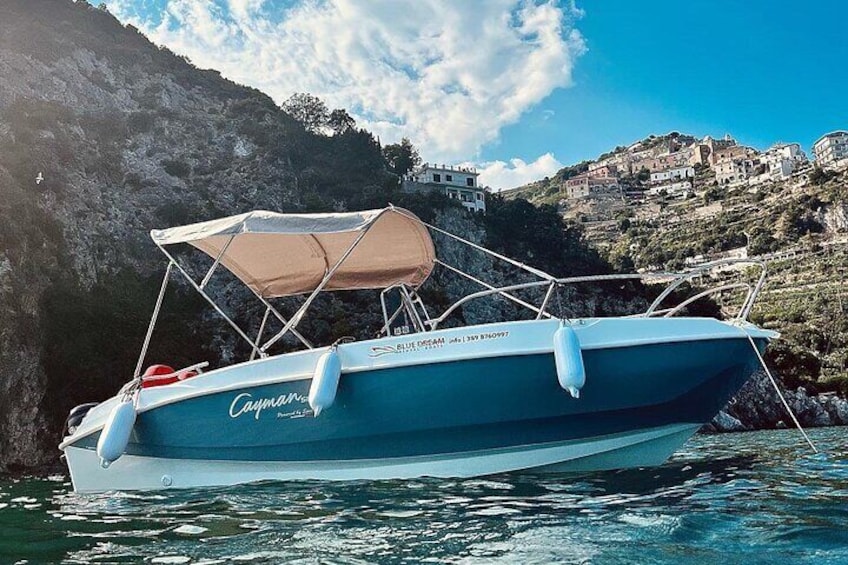 A boat named 'Cayman' with a canopy, anchored near a rocky coastline with buildings in the background. 1