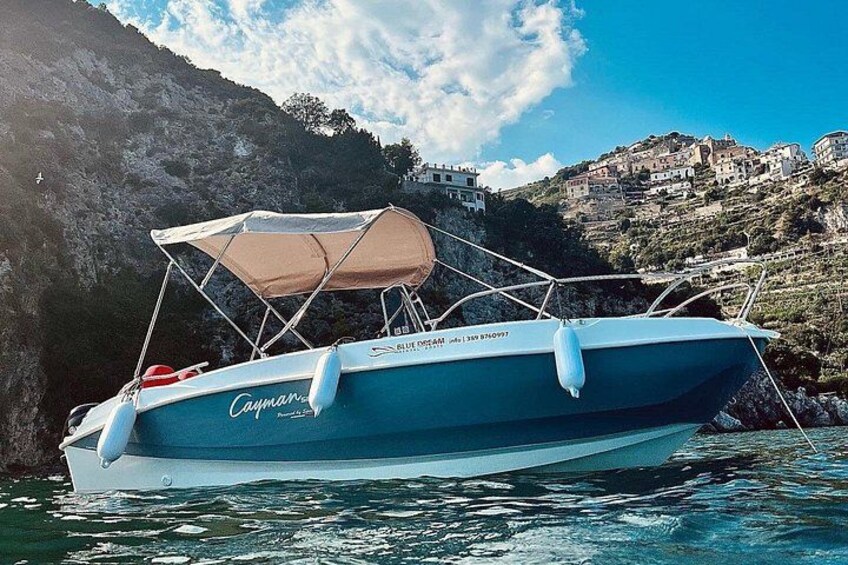 A boat named 'Cayman' with a canopy, anchored near a rocky coastline with buildings in the background. 1