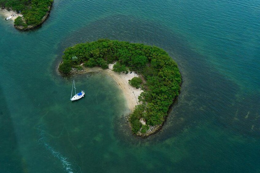 Vue aérienne d’une petite île couverte d’une végétation luxuriante et verdoyante, bordée d’eaux turquoise et cristallines. Un voilier est amarré près du rivage. 3