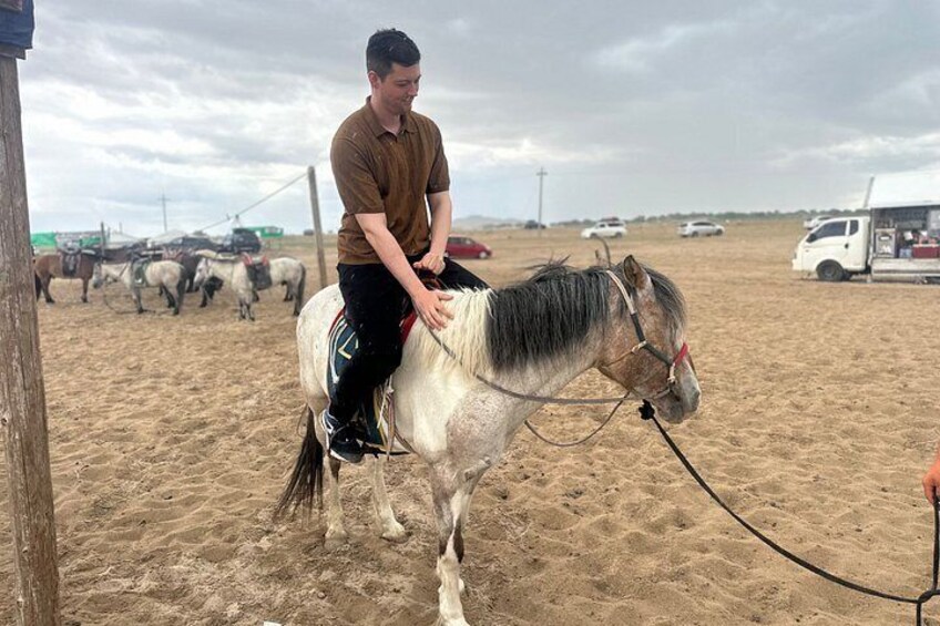 A person riding a white horse with a black mane on a sandy beach, with other horses and vehicles in the background. 2