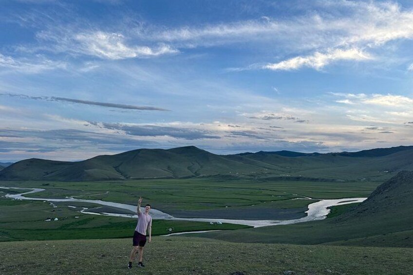 A person standing on a grassy hill overlooking a river and distant mountains under a partly cloudy sky. 4