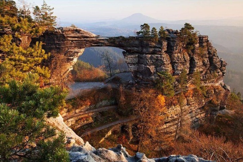 Una scogliera rocciosa con un arco naturale, circondata da alberi e una catena montuosa in lontananza. 1