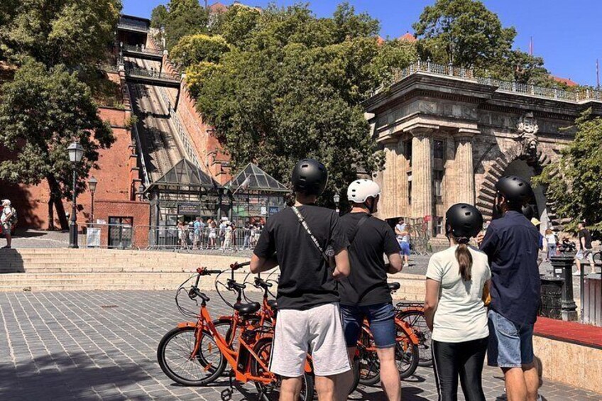 A group of people standing near bicycles, with a large building and trees in the background. 4