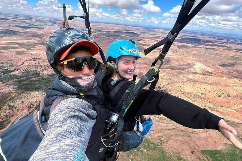 Two people on a tandem paragliding flight with a vast landscape below. 1