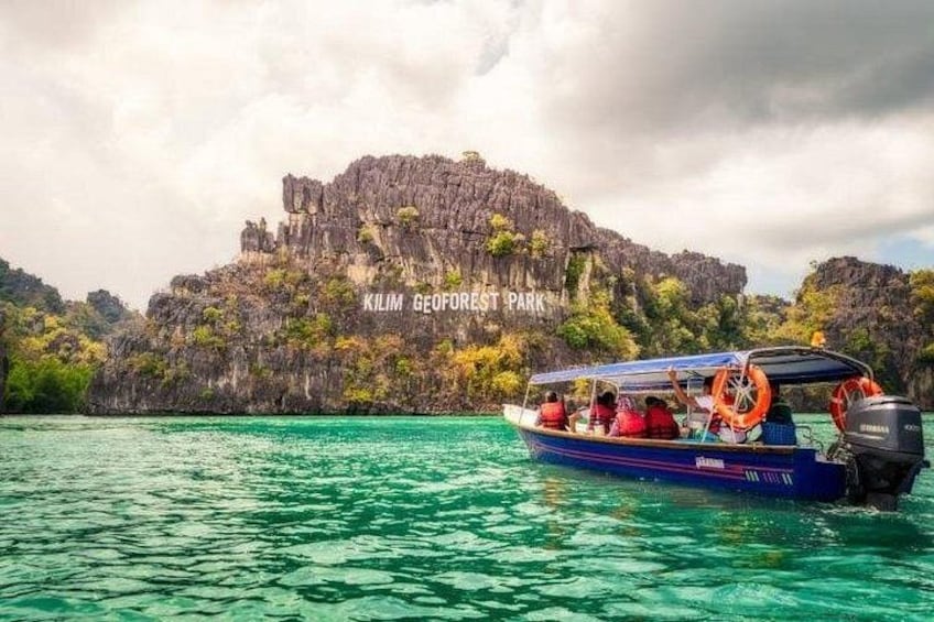 Ein Boot auf türkisblauem Wasser mit einer steinigen Klippe im Hintergrund, auf der 'Kilim GeoForest Park' steht. 2