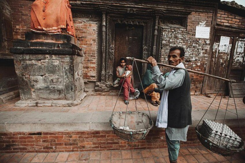 A man balancing a long pole with baskets on either end, standing in front of a traditional building with a statue on a pedestal. 5
