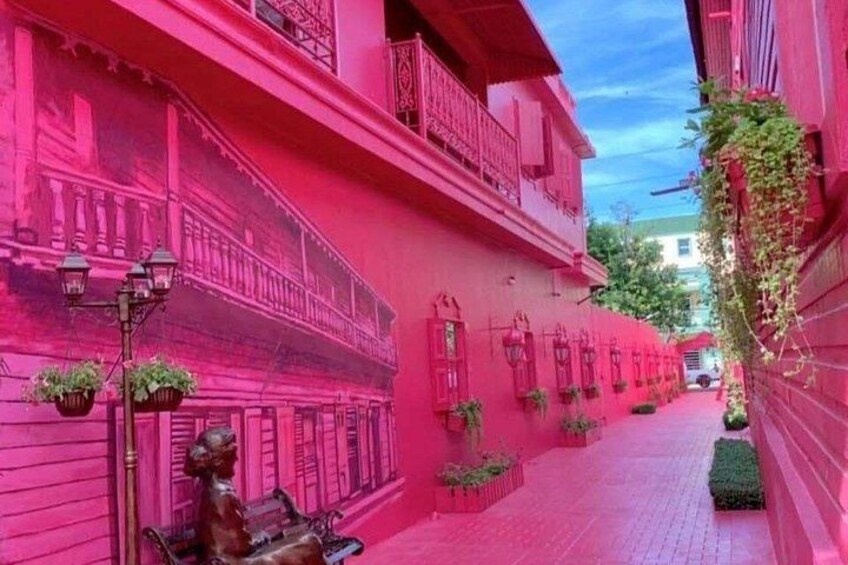 Une rue étroite bordée de bâtiments roses, avec une statue et des plantes suspendues. 5