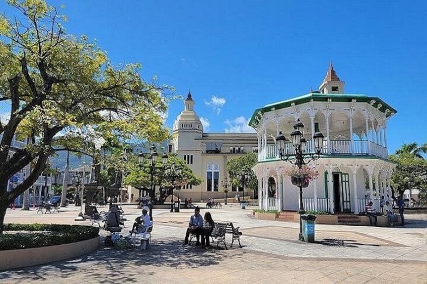 Une place avec un kiosque, un bâtiment surmonté d’un dôme et des personnes assises sur des bancs. 1