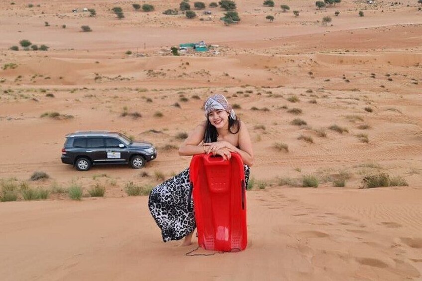 A person in a patterned skirt and headscarf sitting on a sand dune with a red bag, a black SUV in the background, and sparse vegetation. 1