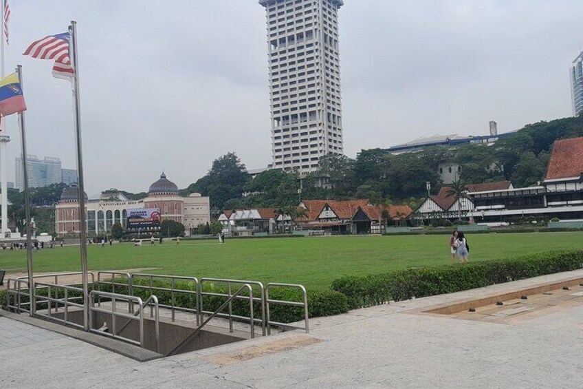 Un parque con un amplio césped, un asta con banderas, un edificio con cúpula y un rascacielos alto al fondo. 2