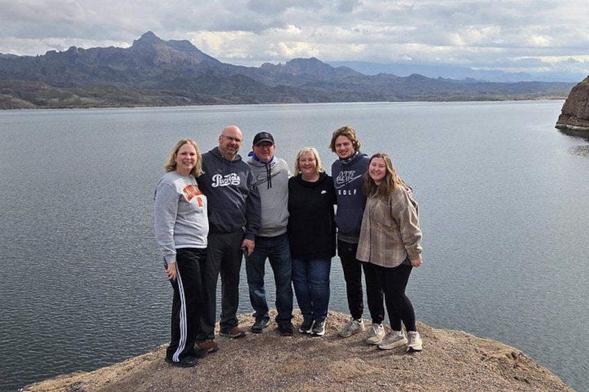 A group of people standing on a rocky outcrop by a lake with mountains in the background. 4