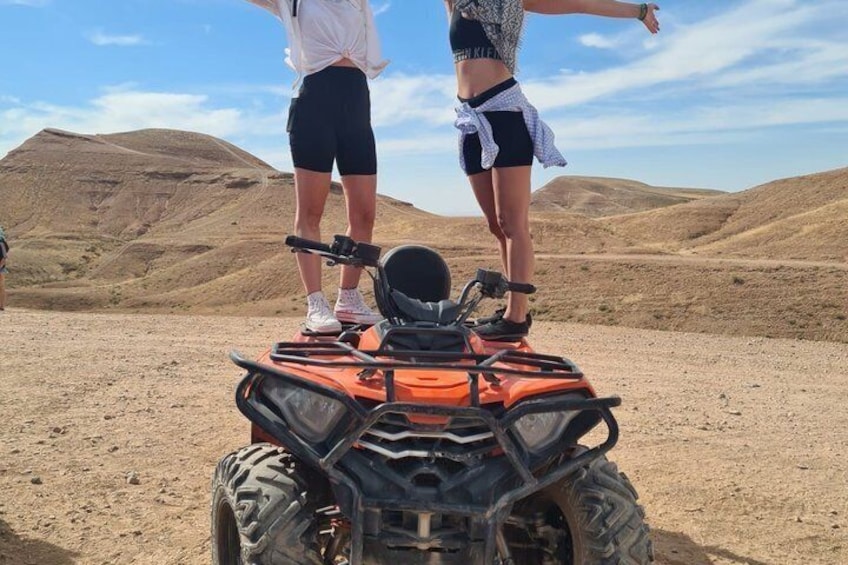 Two people standing on an orange ATV in a desert landscape with hills in the background. 1