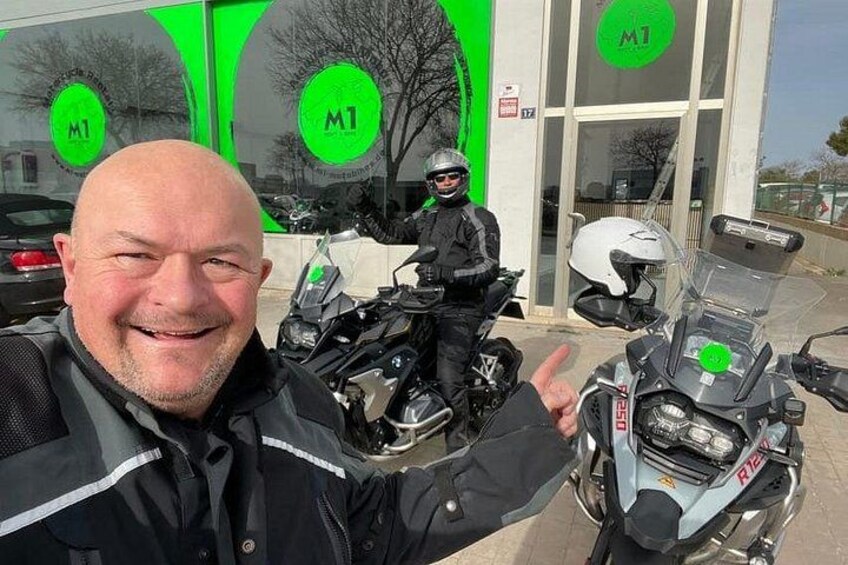 Un homme vêtu d’une veste noire et d’un casque, une moto et un bâtiment avec une enseigne verte. 1
